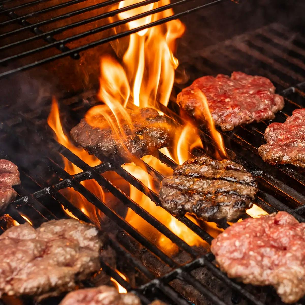 Burgers cooking on a flaming charcoal grill.