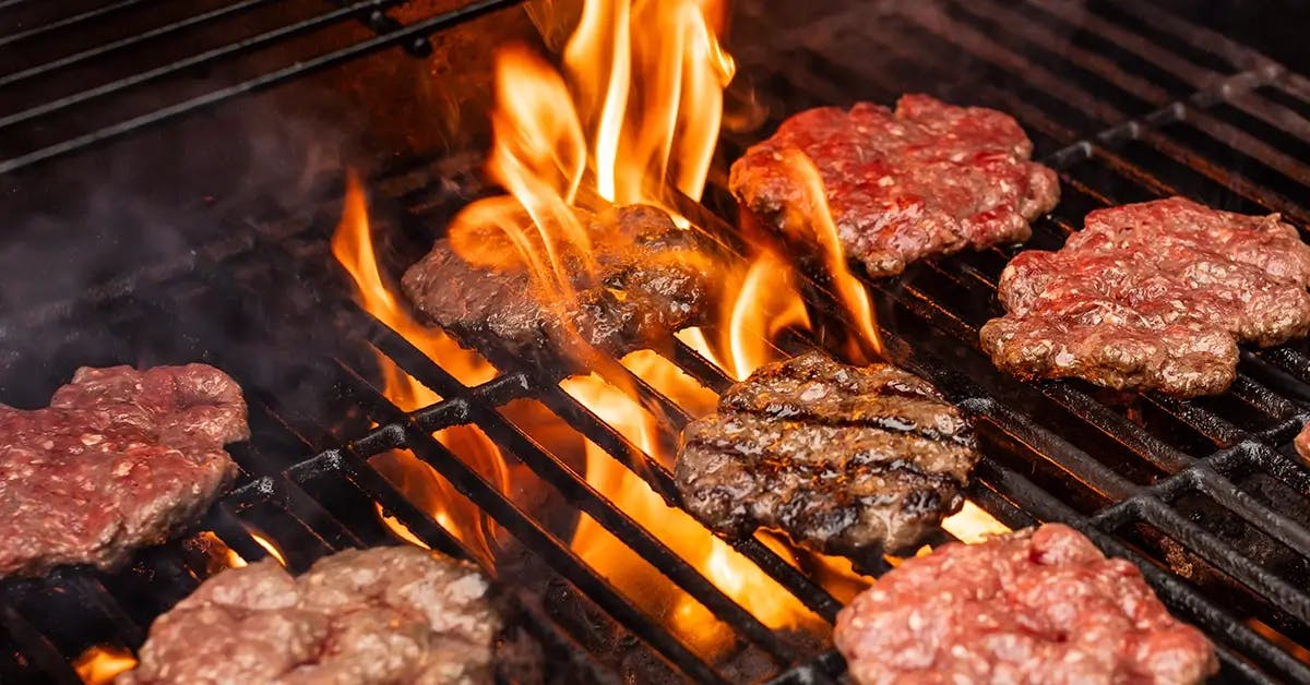 Burgers cooking on a flaming charcoal grill.